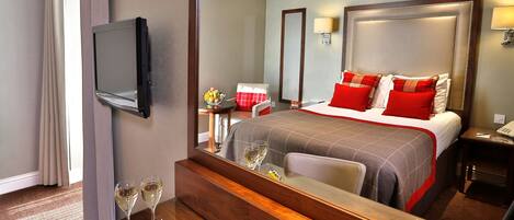 A hotel room with a large bed, a bedside table with a bowl of fruit and glasses, a TV mounted on the wall, and a mirror reflecting the room.