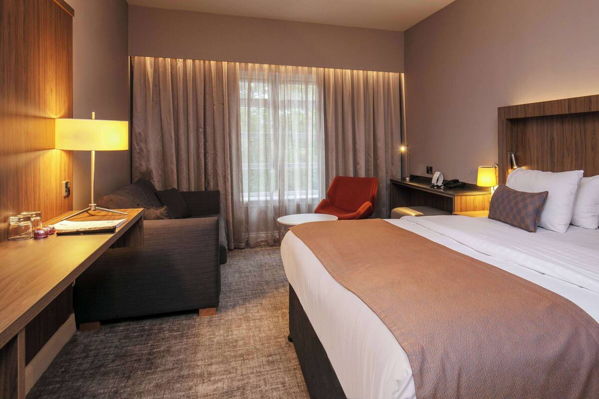 A hotel room with a bed, a desk, a chair, a small table, and a window with curtains.