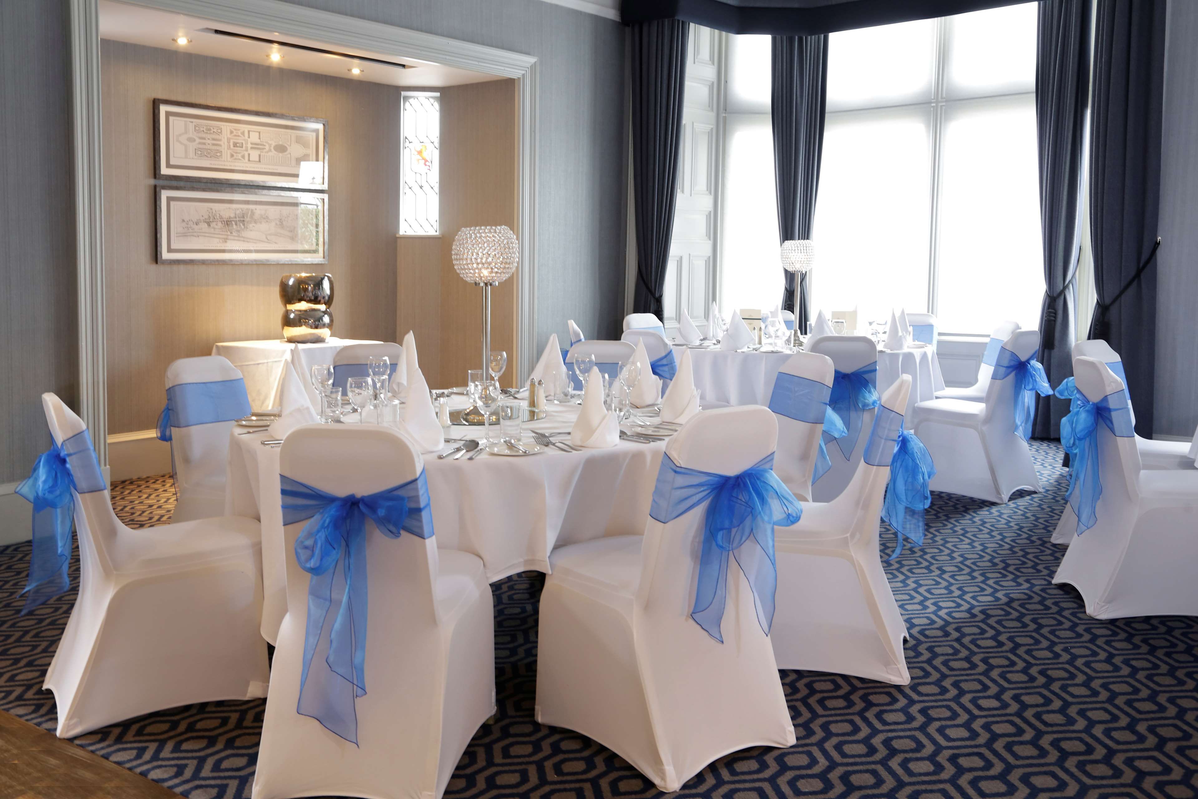 A banquet hall with round tables, white chairs with blue sashes, and a patterned carpet.