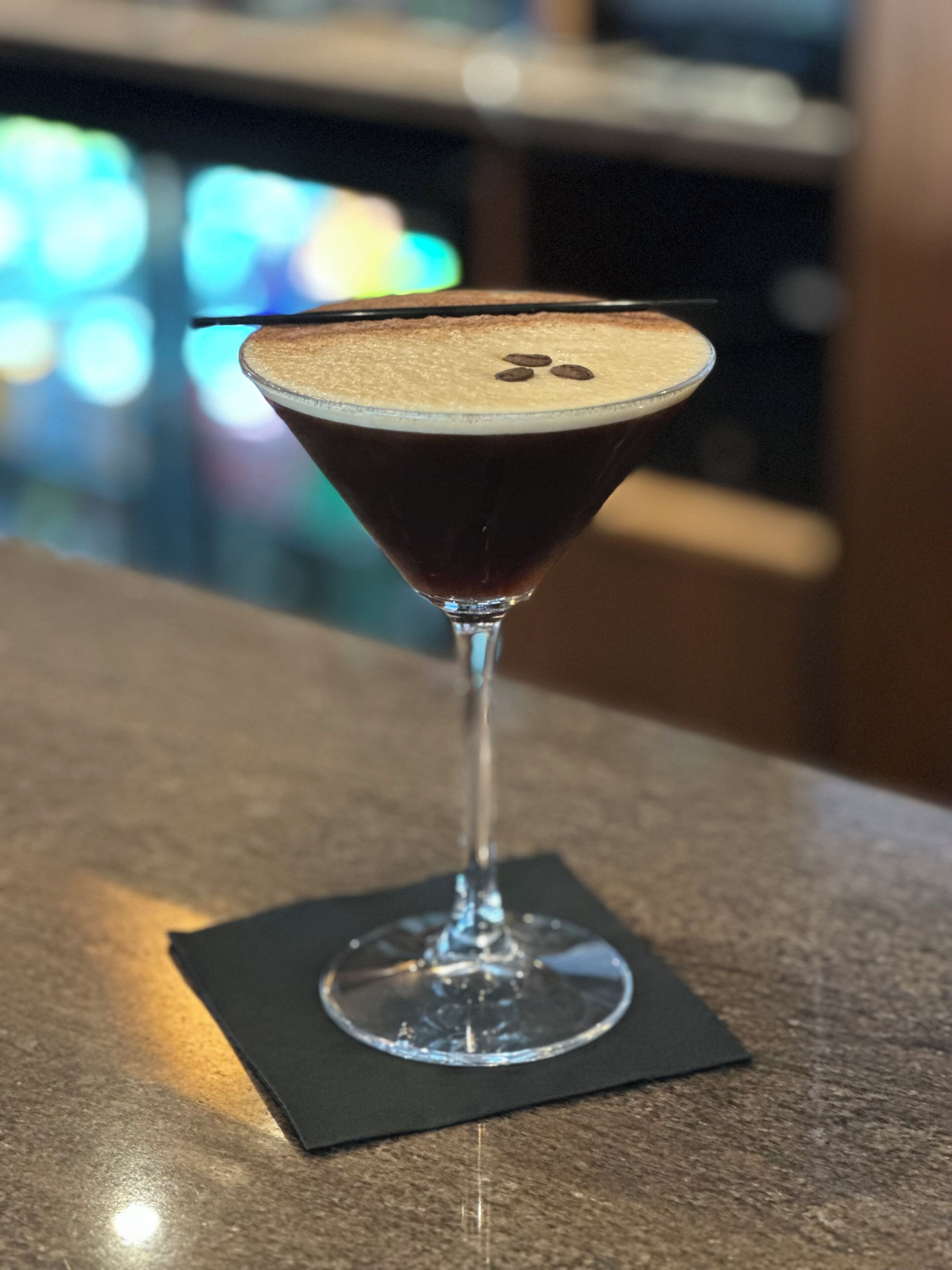 Ein Martini-Glas mit Schaumkrone auf einem dunklen Untersetzer, platziert auf einer strukturierten Oberfläche.