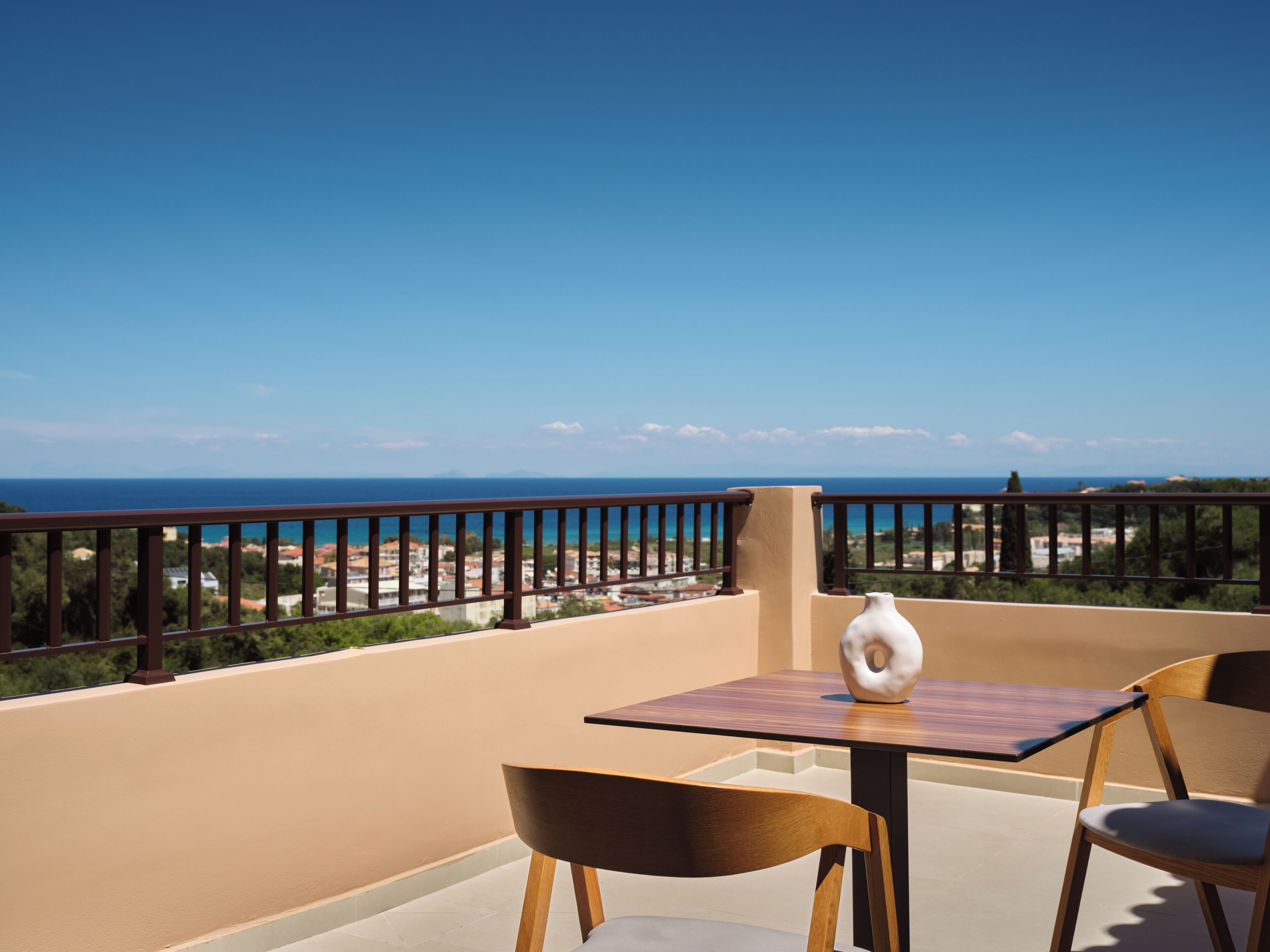 A balcony with a table and chairs, a vase, and a view of the sea and distant landscape.