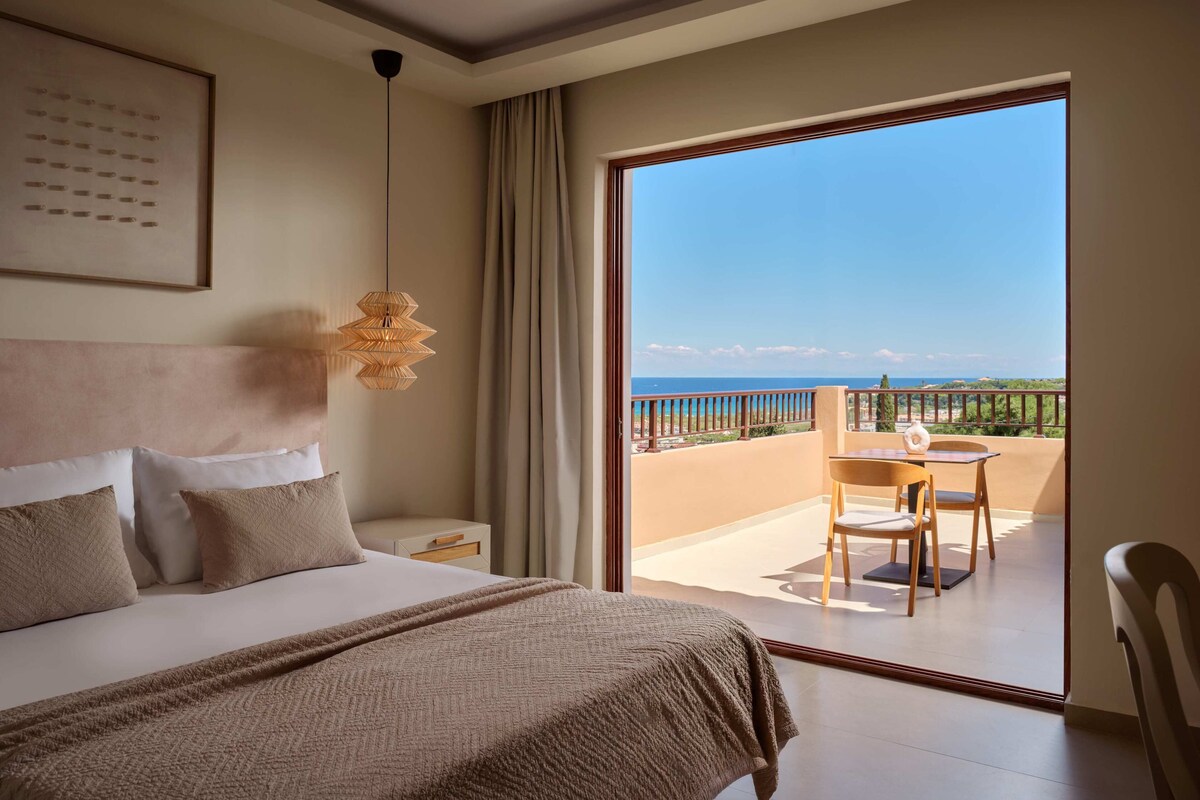 A hotel room with a bed, a bedside table, a hanging light fixture, a balcony with a table and chair, and a view of the sea.