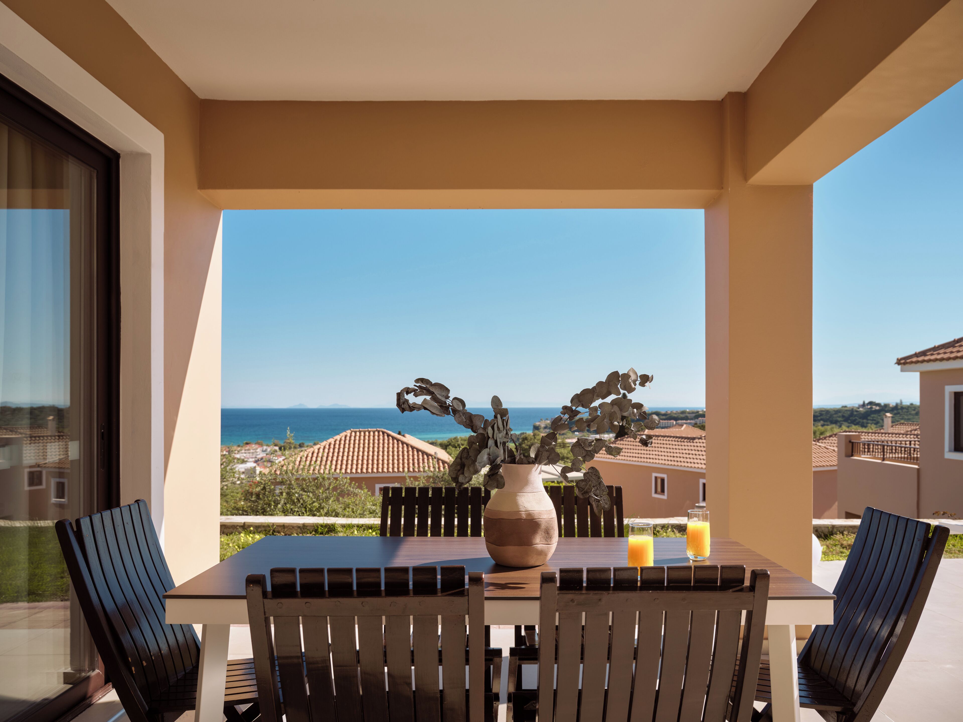 A balcony with a table, chairs, a vase with plants, and a view of the sea and buildings.