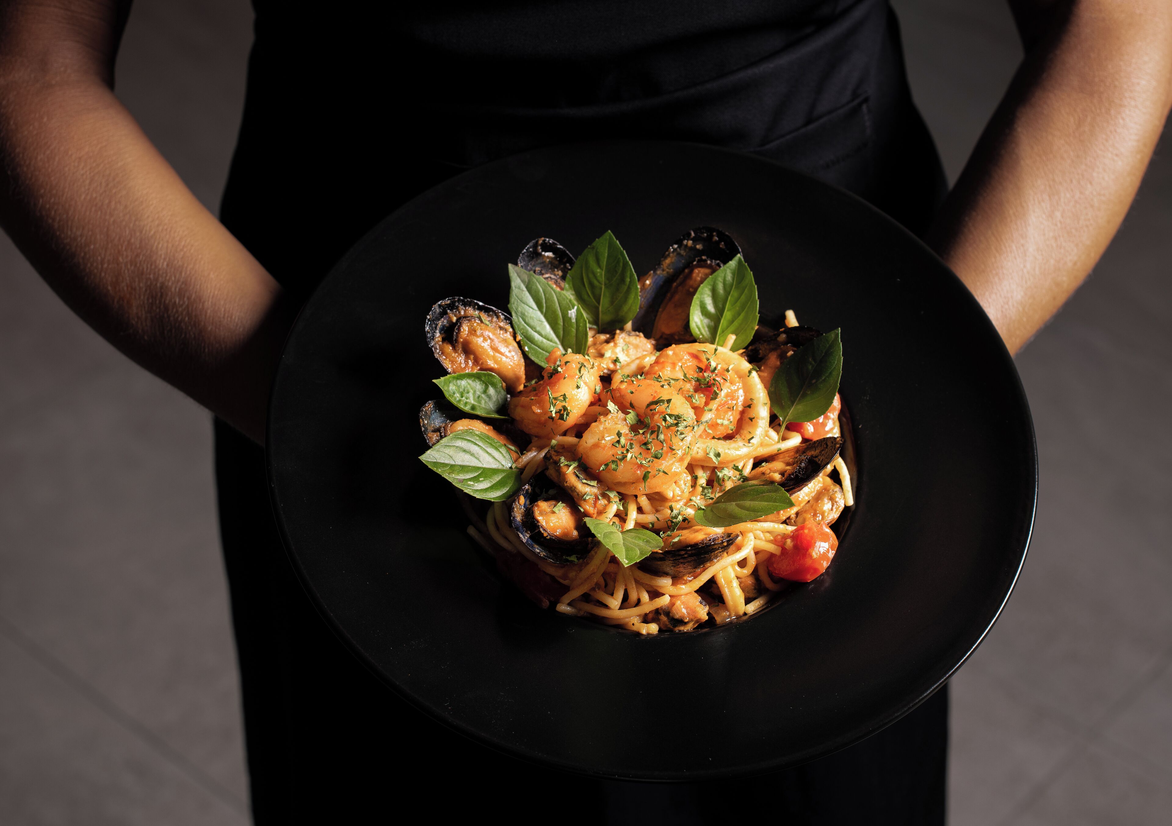 A person holding a black plate with a pasta dish, garnished with basil leaves.