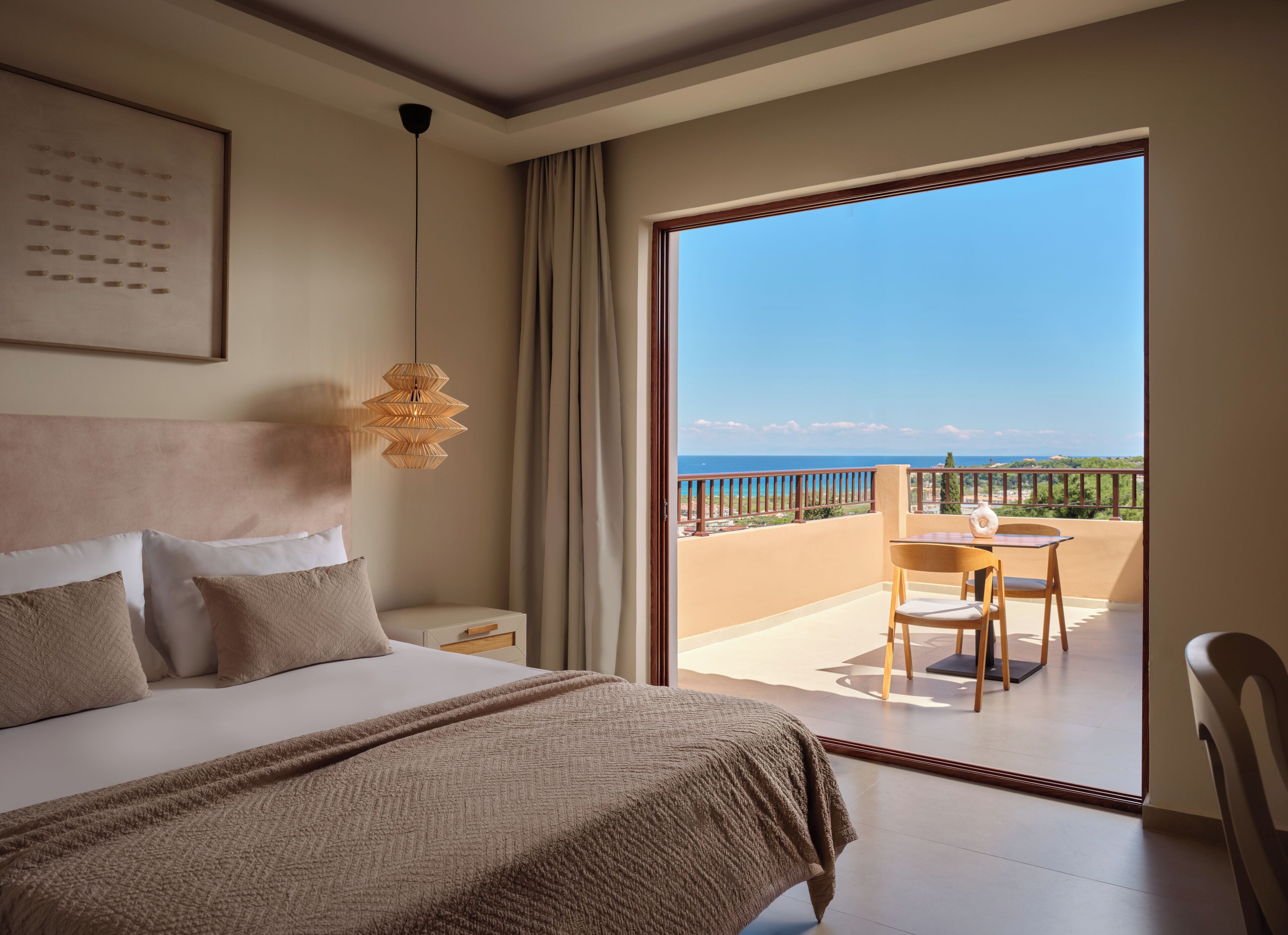 Ein Schlafzimmer mit einem großen Bett, einem Nachttisch, einer Hängeleuchte und Blick auf einen Balkon mit Tisch und Stuhl.