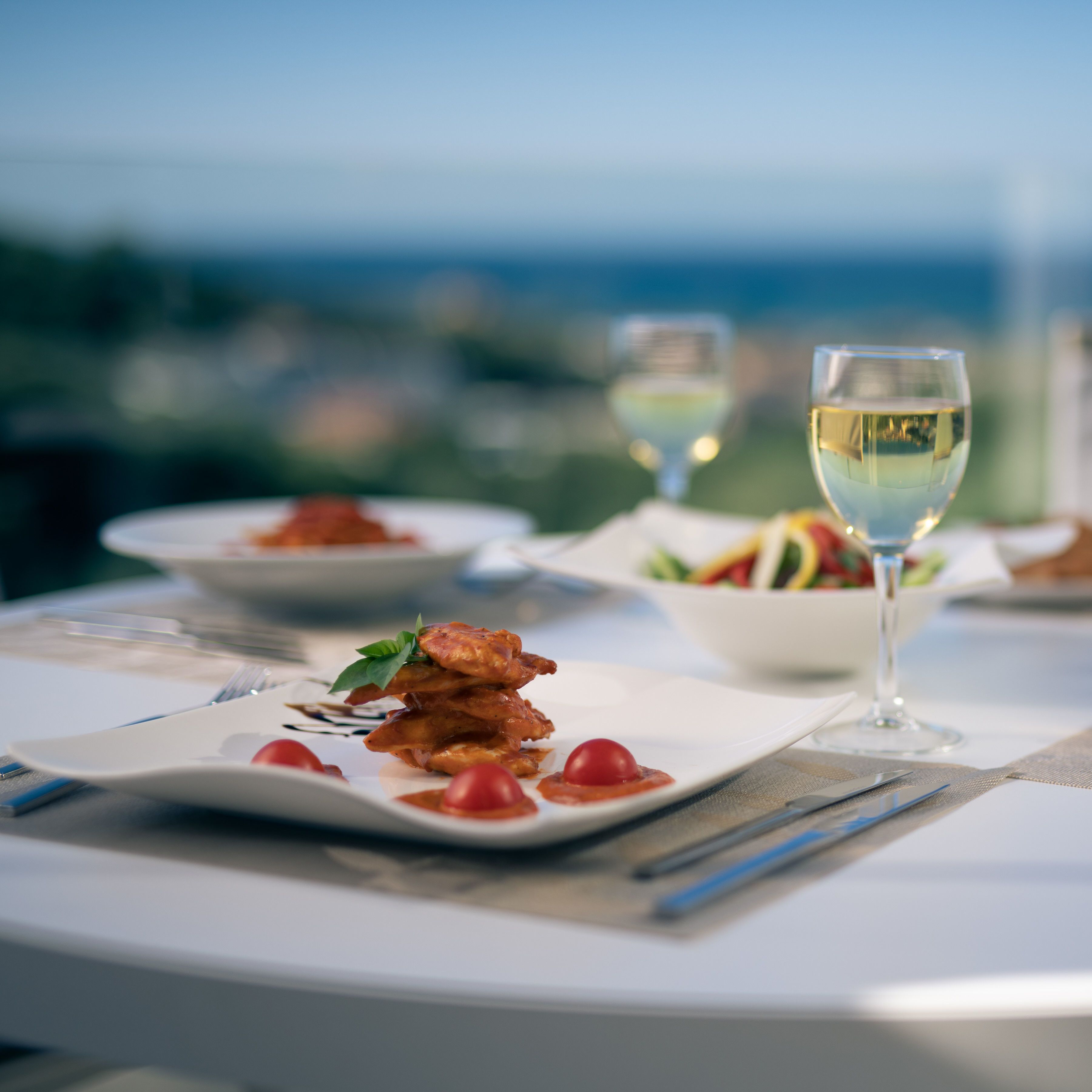 Una mesa de comedor con platos de comida, una copa de vino blanco y vistas al exterior.