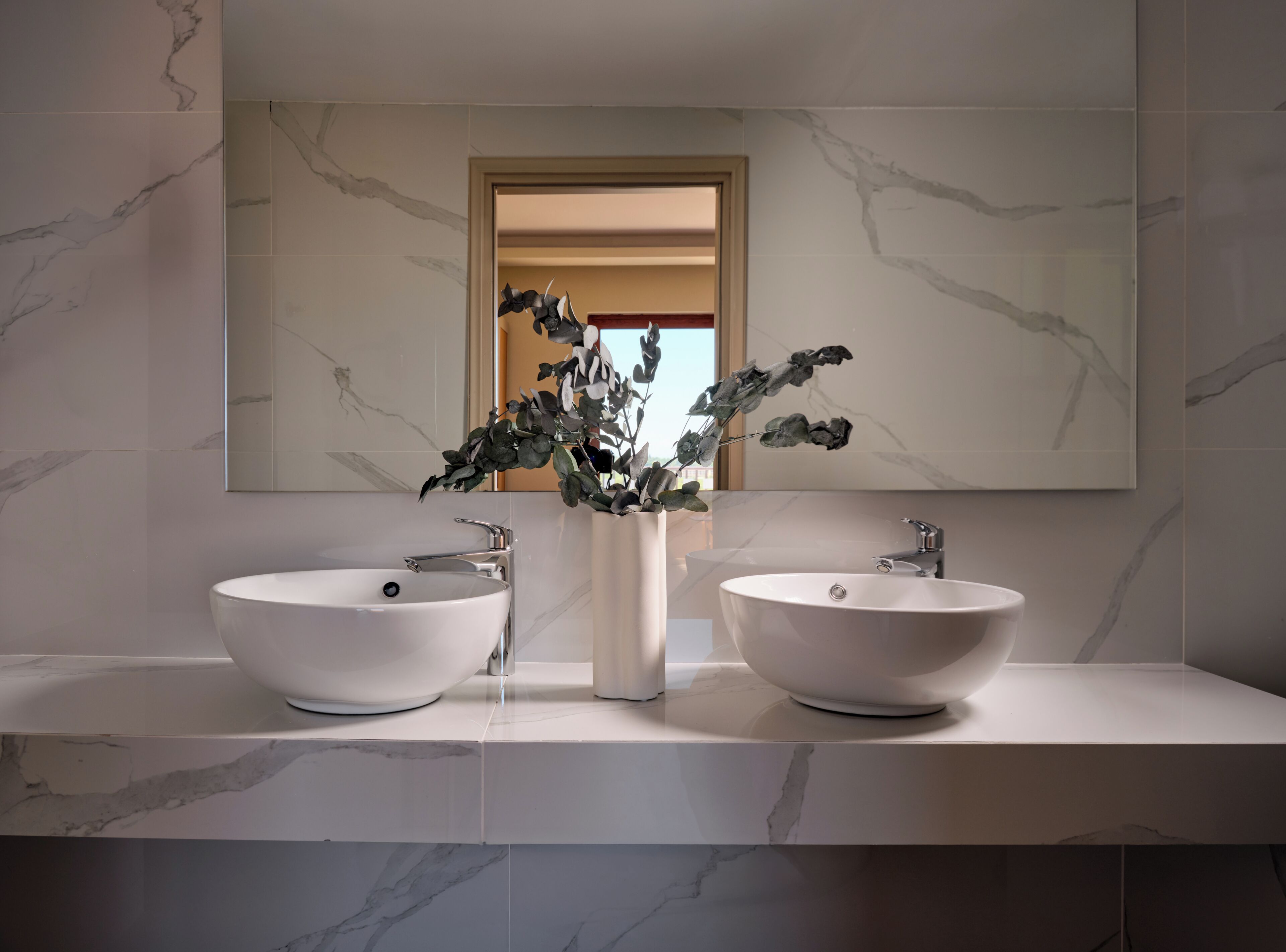 A modern bathroom with two white vessel sinks, a large mirror, and a vase with dried flowers.