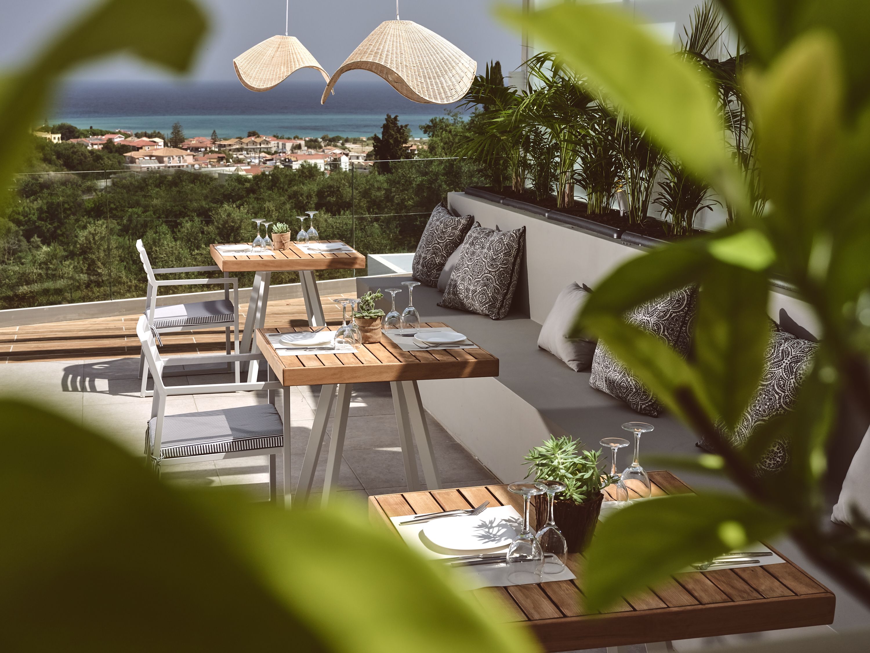 Un área de comedor al aire libre con mesas y sillas de madera, rodeada de plantas en macetas y cojines estampados.