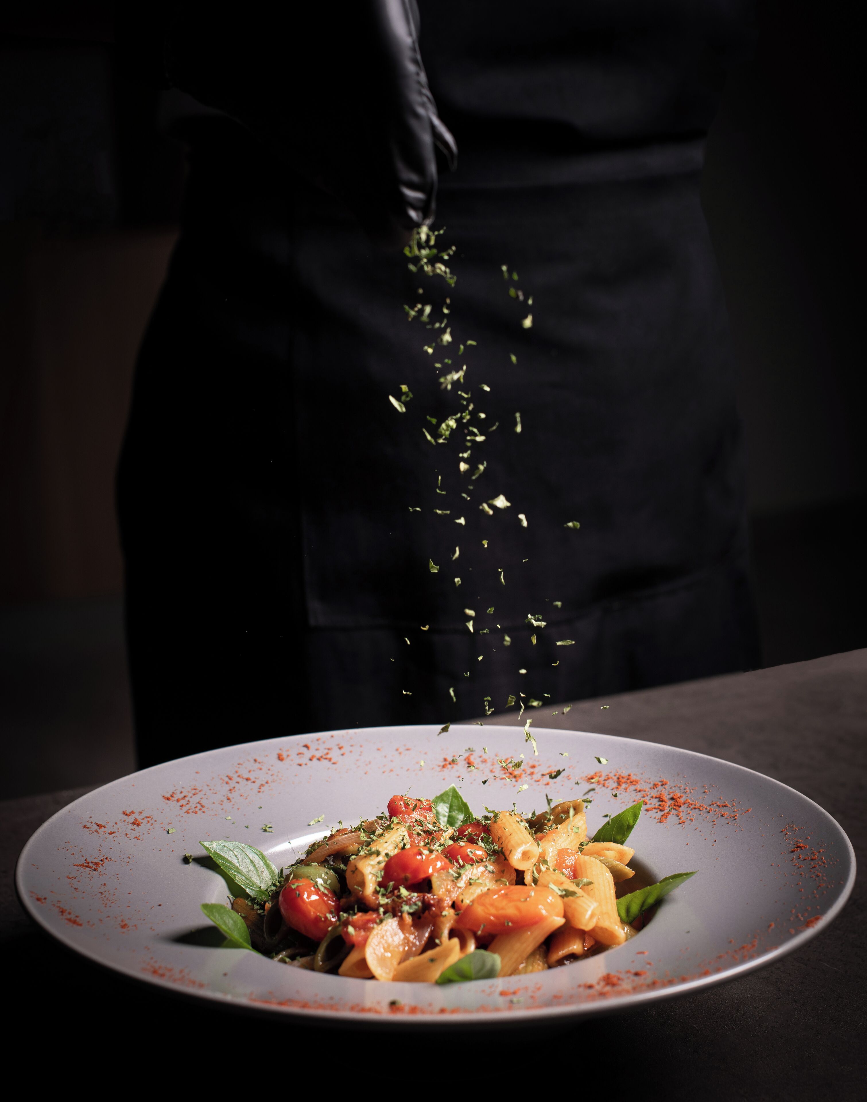 A chef sprinkling herbs over a dish of pasta and vegetables.