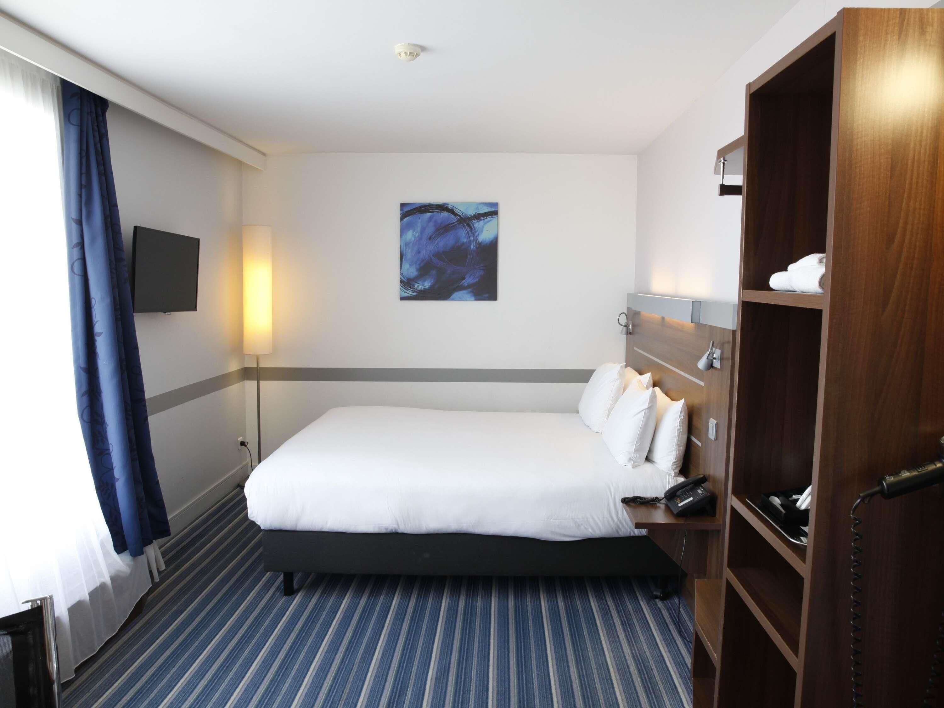 Habitación de hotel con cama, mesita de noche con teléfono, televisor montado en la pared y un cuadro.