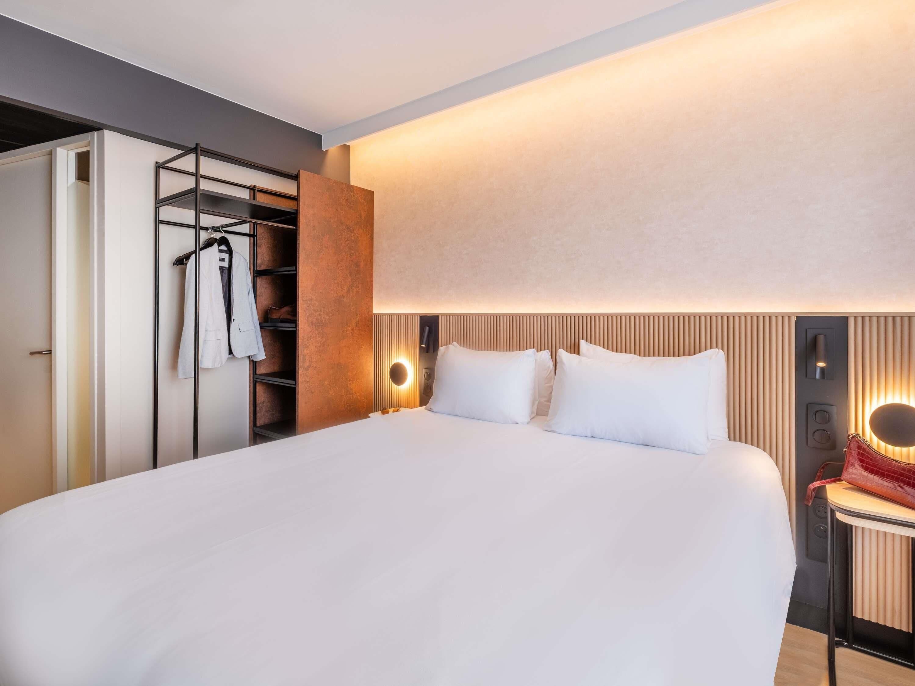 Une chambre d’hôtel moderne, dotée d’un grand lit, d’une armoire où sont suspendus des vêtements et d’une table de chevet avec une lampe.