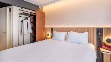 Una habitación de hotel moderna con una cama grande, un armario con ropa colgada y una mesita de noche con una lámpara.