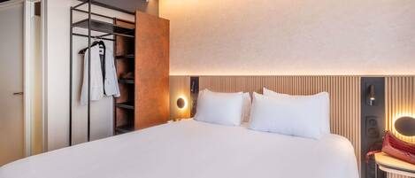 Une chambre d’hôtel moderne, dotée d’un grand lit, d’une armoire où sont suspendus des vêtements et d’une table de chevet avec une lampe.