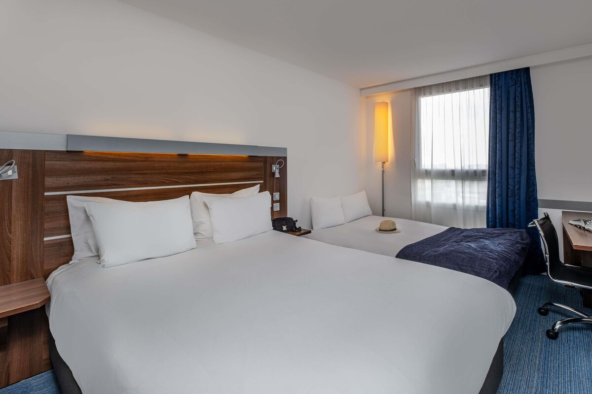 Une chambre d’hôtel avec deux lits, un bureau, une chaise, une lampe et une fenêtre avec des rideaux.