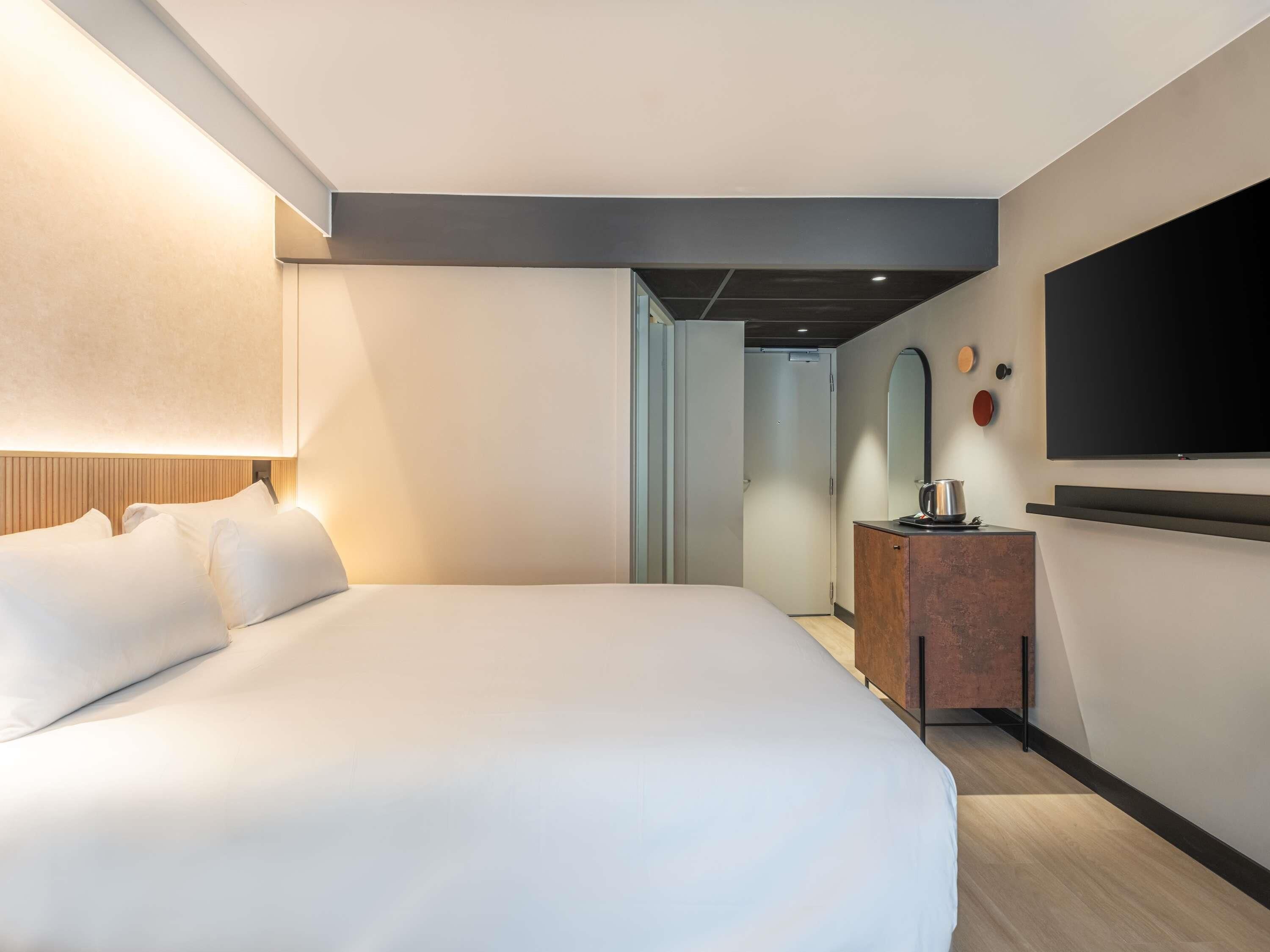 A modern hotel room with a large bed, a flat-screen TV, a wooden dresser, and a mirror.
