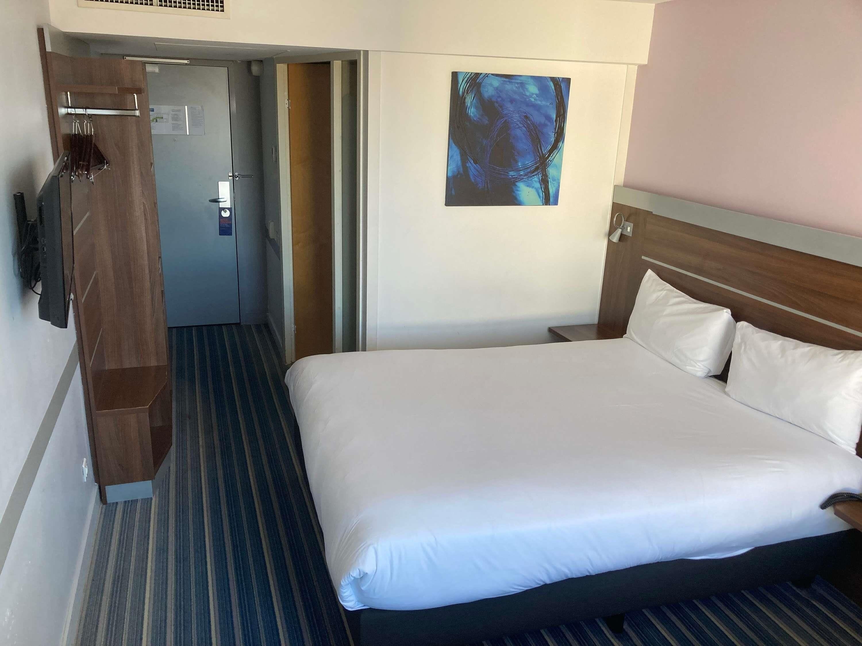 A hotel room with a large bed, a wooden wardrobe, a painting on the wall, and a door leading to another room.