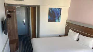 A hotel room with a large bed, a wooden wardrobe, a painting on the wall, and a door leading to another room.