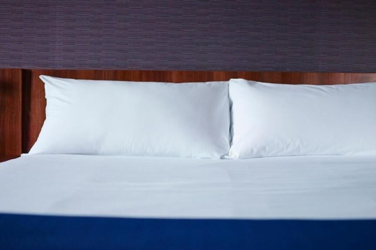 Una cama bien hecha con ropa blanca y falda azul, frente a un cabecero de madera.