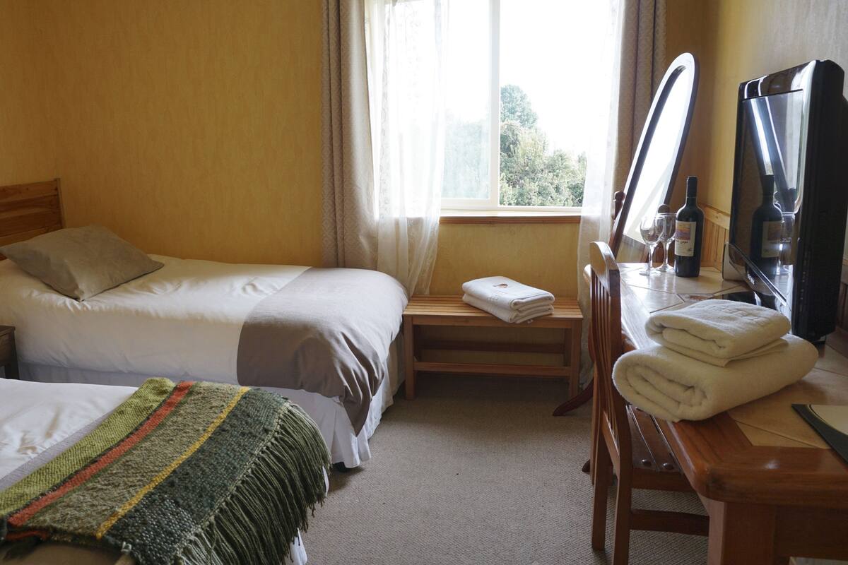A hotel room with two beds, a wooden nightstand with a lamp, a wooden bench, a desk with a mirror, and a TV.