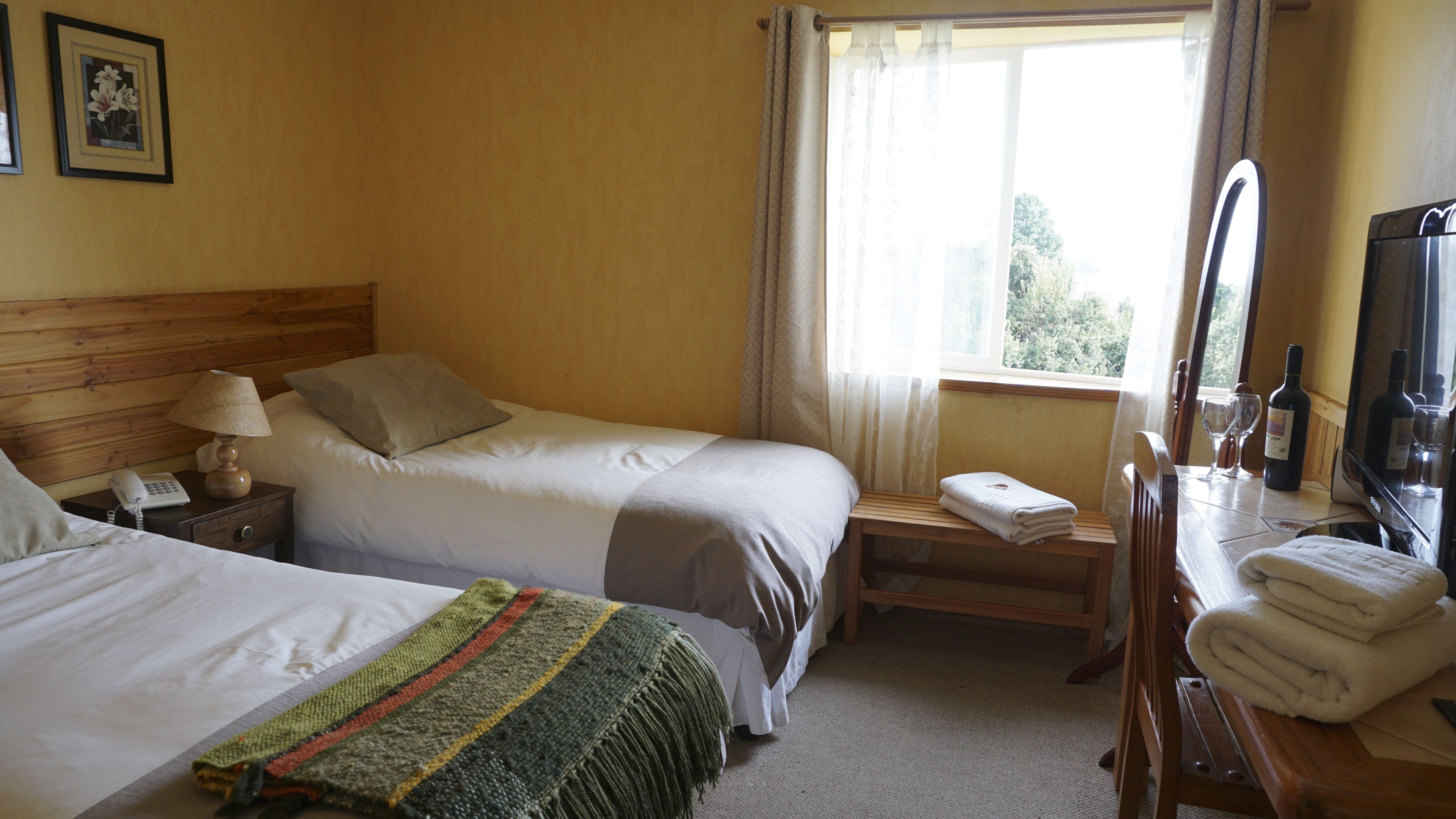 A hotel room with two beds, a wooden headboard, a bedside table with a lamp, a small table with a towel, a mirror, and a window with curtains.