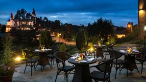 An outdoor dining area with round tables and chairs, surrounded by potted plants and a view of a castle in the background. - Solace Hotel Puerto Varas (Puerto Varas)