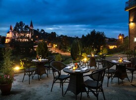 Un espacio de comedor al aire libre con mesas y sillas redondas, rodeado de plantas en macetas y una vista de un castillo al fondo.