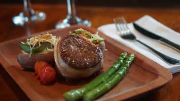 Een houten tafel met een bord eten, waaronder een steak, asperges en een kleine tomaat, voor een achtergrond met een open haard.