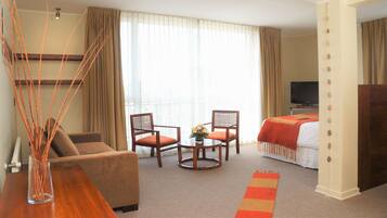 A hotel room with a sofa, two chairs, a small table with a vase, a bed with a red blanket, a TV, and a decorative pillar.