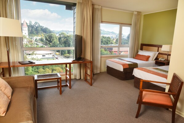Una habitación de hotel con escritorio, dos camas, un sofá y vistas a un pueblo y colinas.