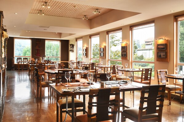 A spacious restaurant with wooden tables and chairs, large windows offering views of greenery, and wall-mounted picture frames. - Solace Hotel Puerto Varas (Puerto Varas)