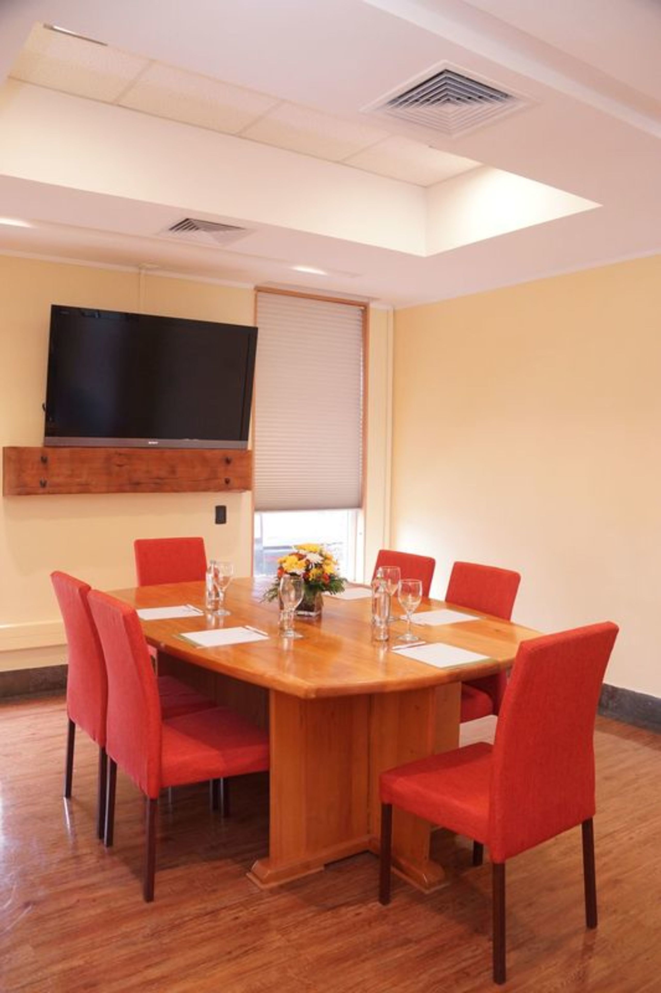 Un salón de conferencias con una mesa de madera, sillas rojas, un televisor de pantalla plana montado en la pared y un ventanal con persianas.