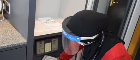 Una persona con uniforme rojo y gorro negro interactuando con un mostrador y una vitrina en una habitación con pared gris y una puerta con pomo dorado.