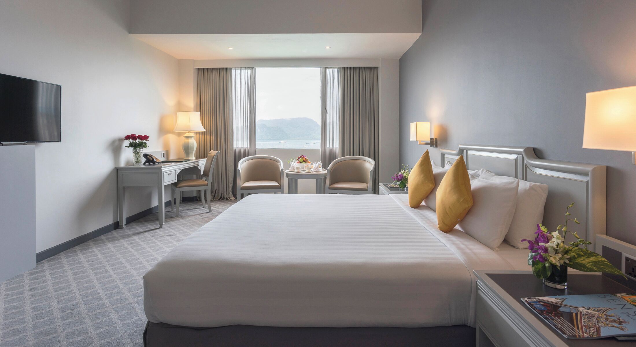 A hotel room with a large bed, a desk, two chairs, a TV, and a window with curtains.
