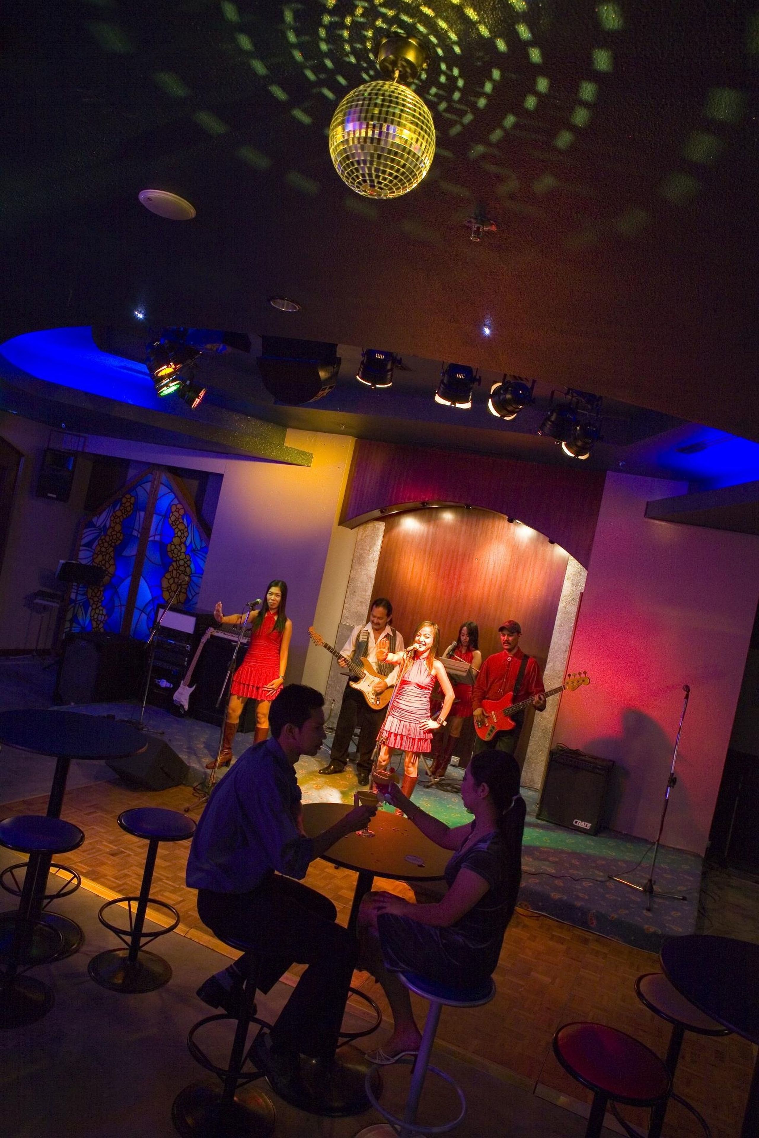 A stage with a band performing, a disco ball, and a bar area with stools.
