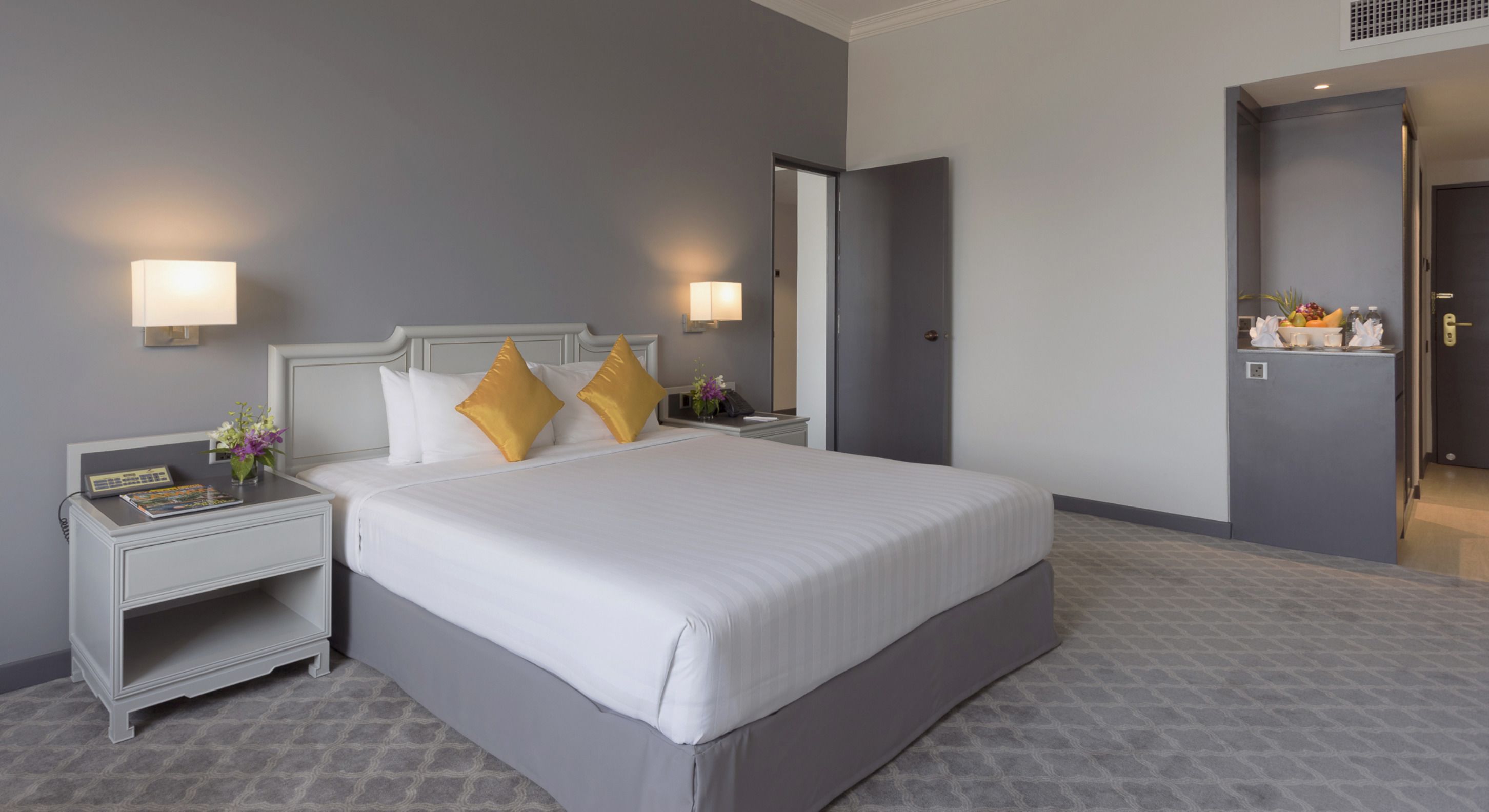 A hotel room with a large bed, two bedside tables with lamps, a nightstand, and a door leading to another room.