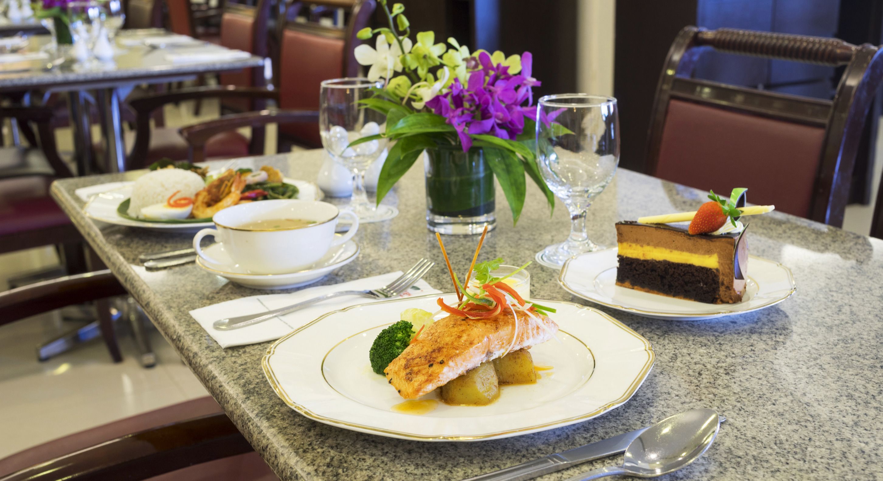 A dining table with plates of food, including salmon, a cake, and a bowl of soup. There are also glasses and a vase with flowers.