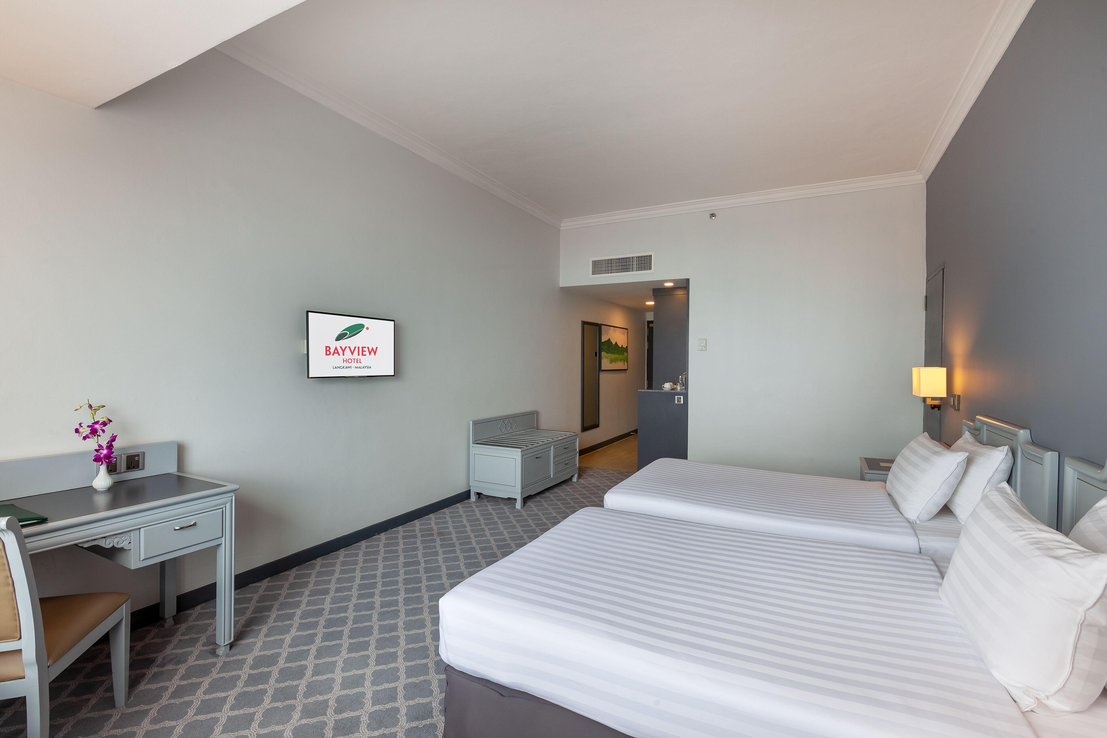 Une chambre d’hôtel avec deux lits, un bureau, une chaise, une petite table ornée d’un vase de fleurs et un panneau encadré accroché au mur.