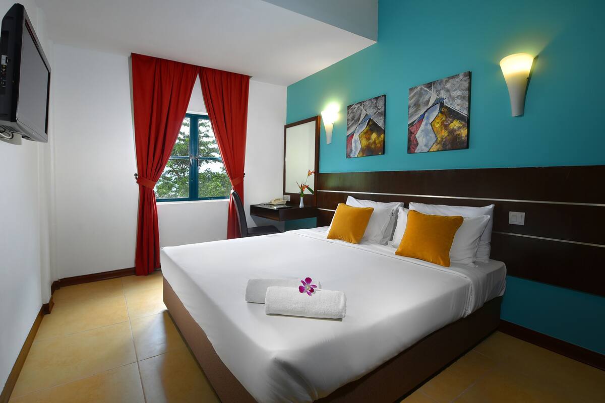 A neatly made bed with white linens and a decorative towel, a nightstand with a lamp, and artwork on the teal-colored wall.