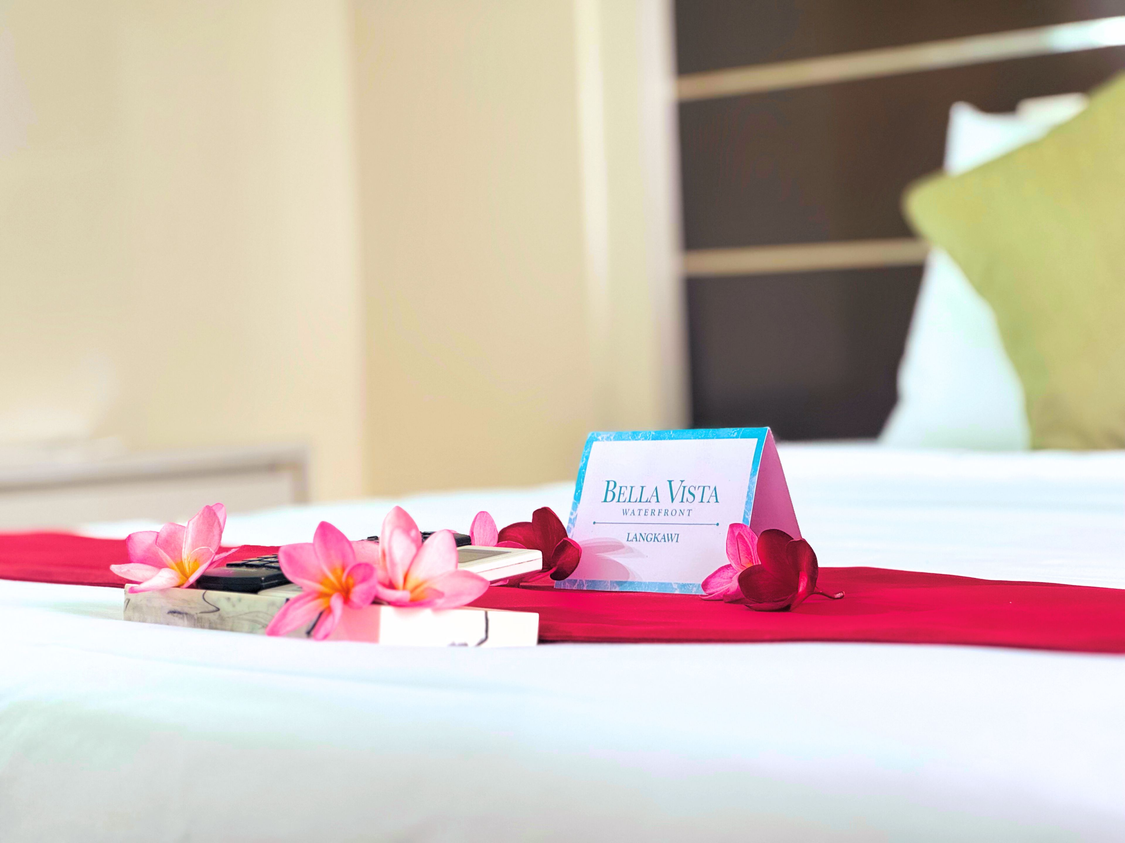A hotel room with a bed, a sign reading 'Bella Vista Waterfront Langkawi,' and a red bedspread.