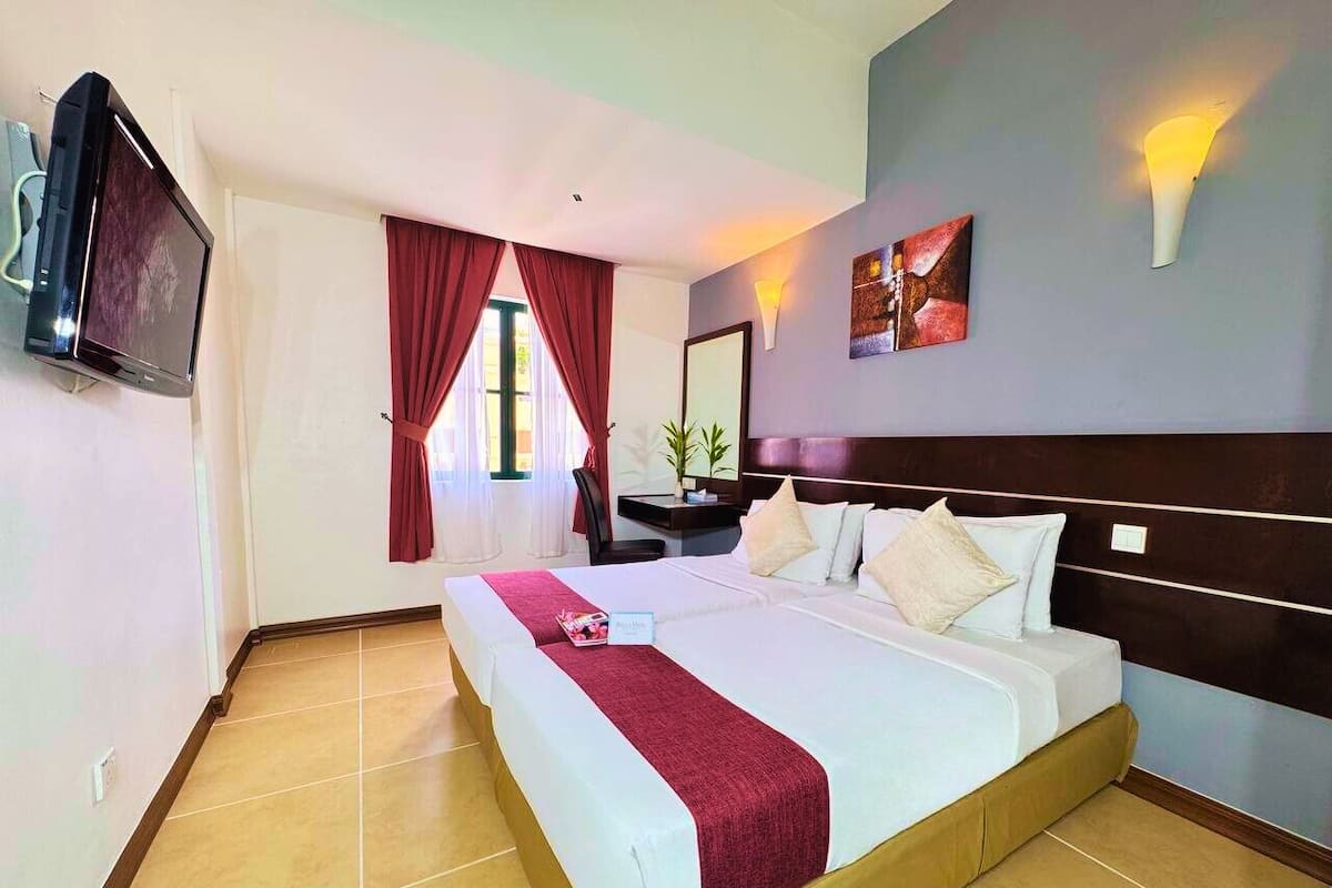 A hotel room with a large bed, a flat-screen TV, a desk with a chair, and a window with red curtains.