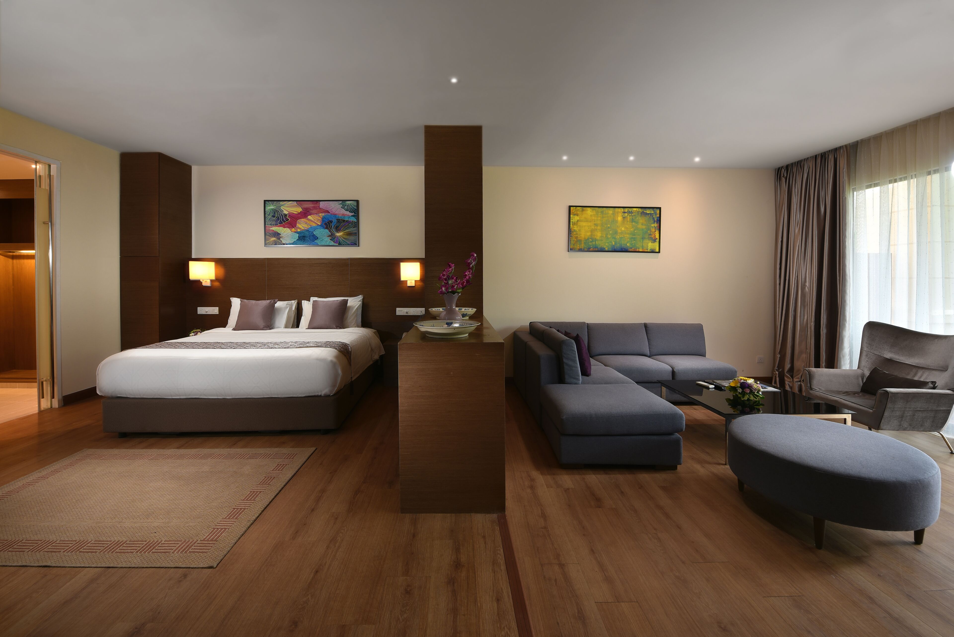 Ett modernt hotellrum med en stor säng, en soffa, ett soffbord och en stol. Det finns två tavlor på väggarna och ett fönster med gardiner.