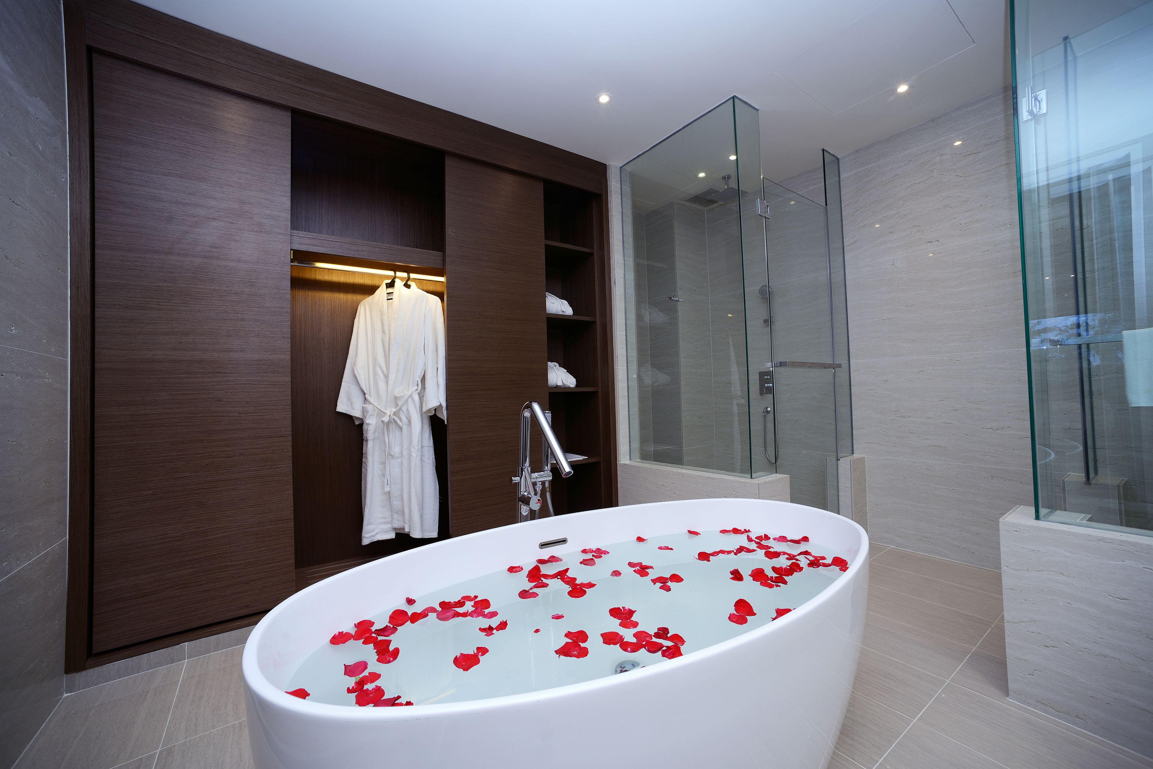 A modern bathroom with a freestanding bathtub filled with rose petals, a glass-enclosed shower, and a walk-in closet with hanging robes.