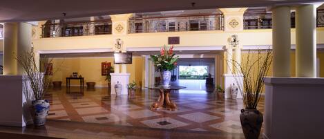 A spacious lobby with a checkered floor, a central table with a vase of flowers, and a balcony with decorative railings.