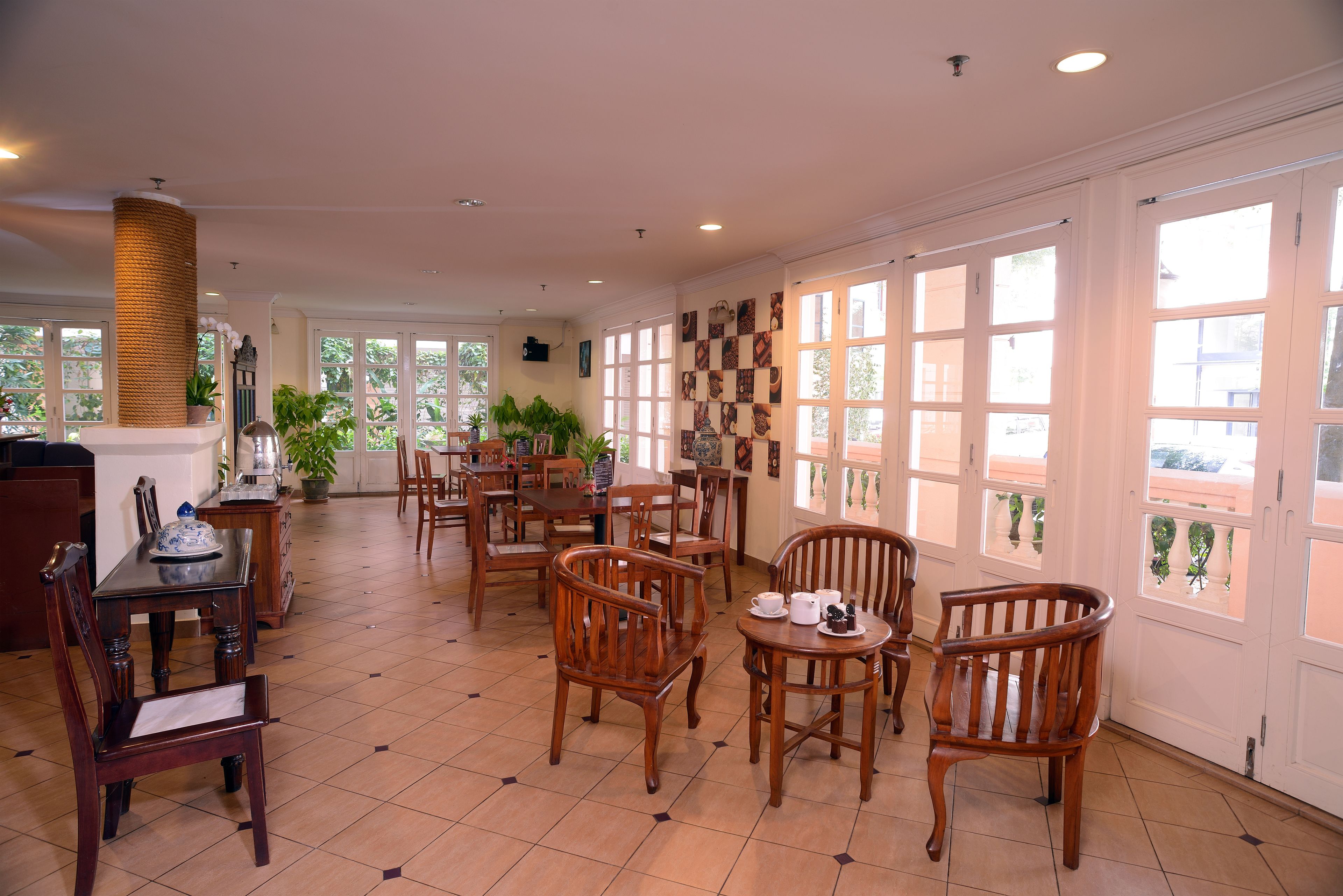 Un comedor amplio con varias mesas y sillas de madera, ventanas grandes con paneles de vidrio decorativos y plantas en macetas.