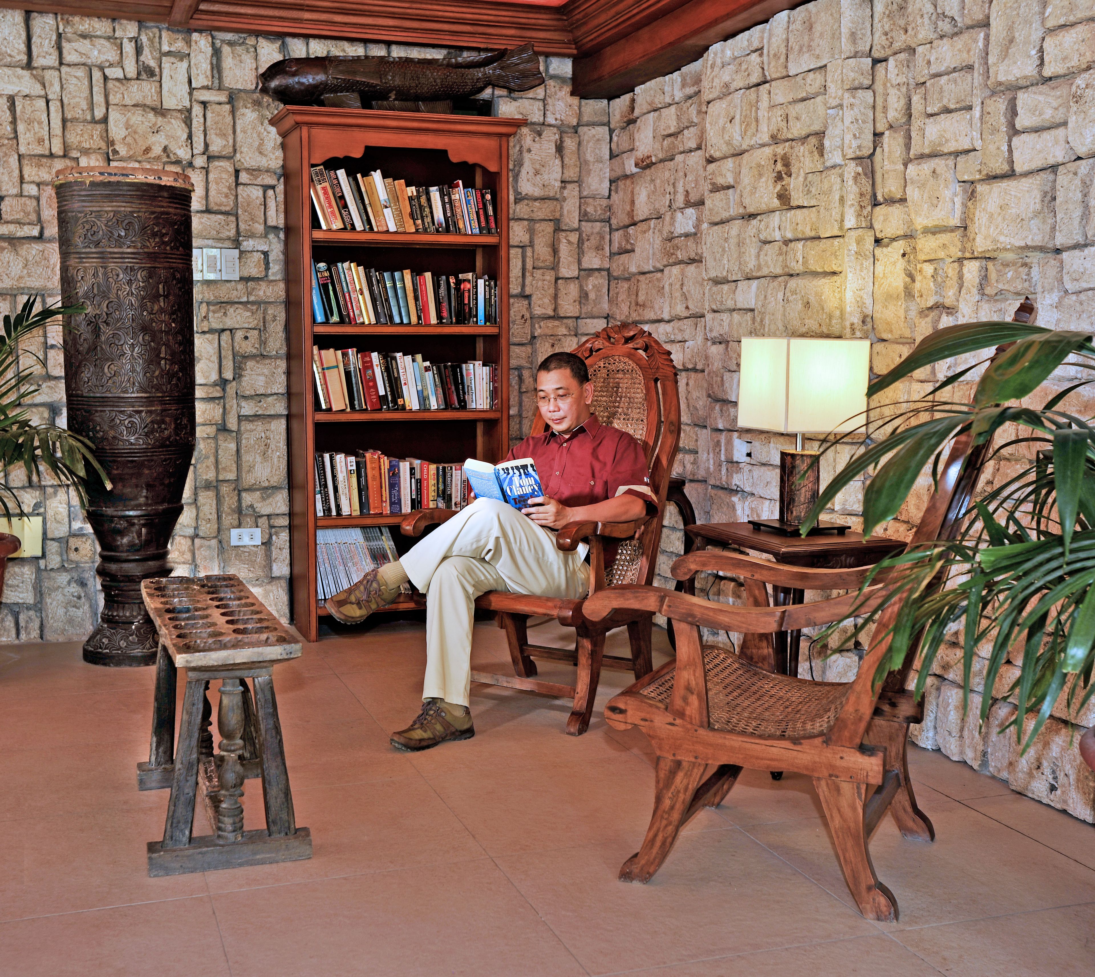 Un hombre leyendo un libro en una habitación con pared de piedra, muebles de madera y una estantería llena de libros.