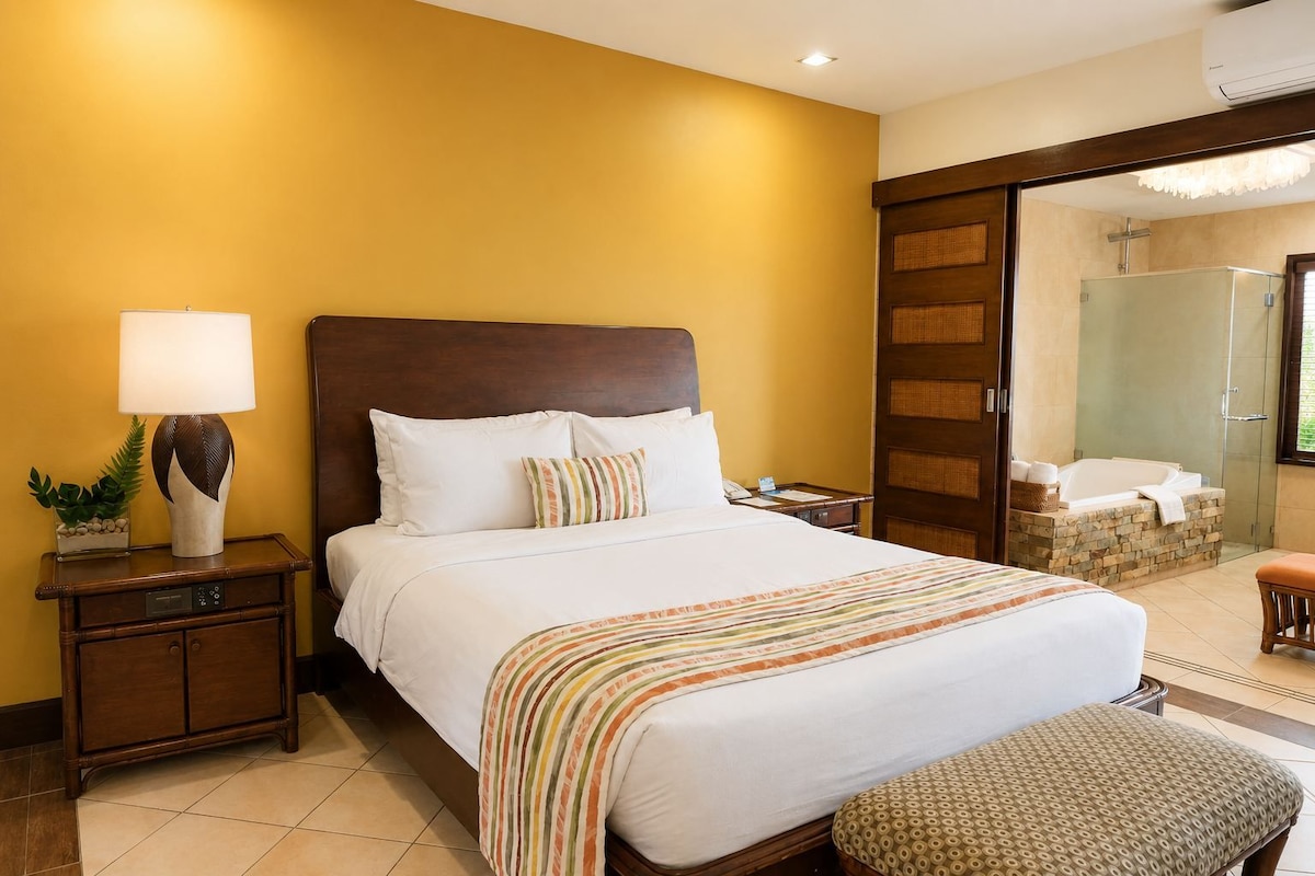 A hotel room with a large bed, a bedside table with a lamp, a small plant, and a bathroom with a bathtub and shower.