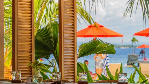Un restaurant situé en bord de mer, avec des meubles en bois, une vue sur l’océan et des parasols rouges.
