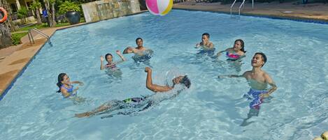 A swimming pool with people enjoying the water, a beach ball, and lounge chairs around the pool.