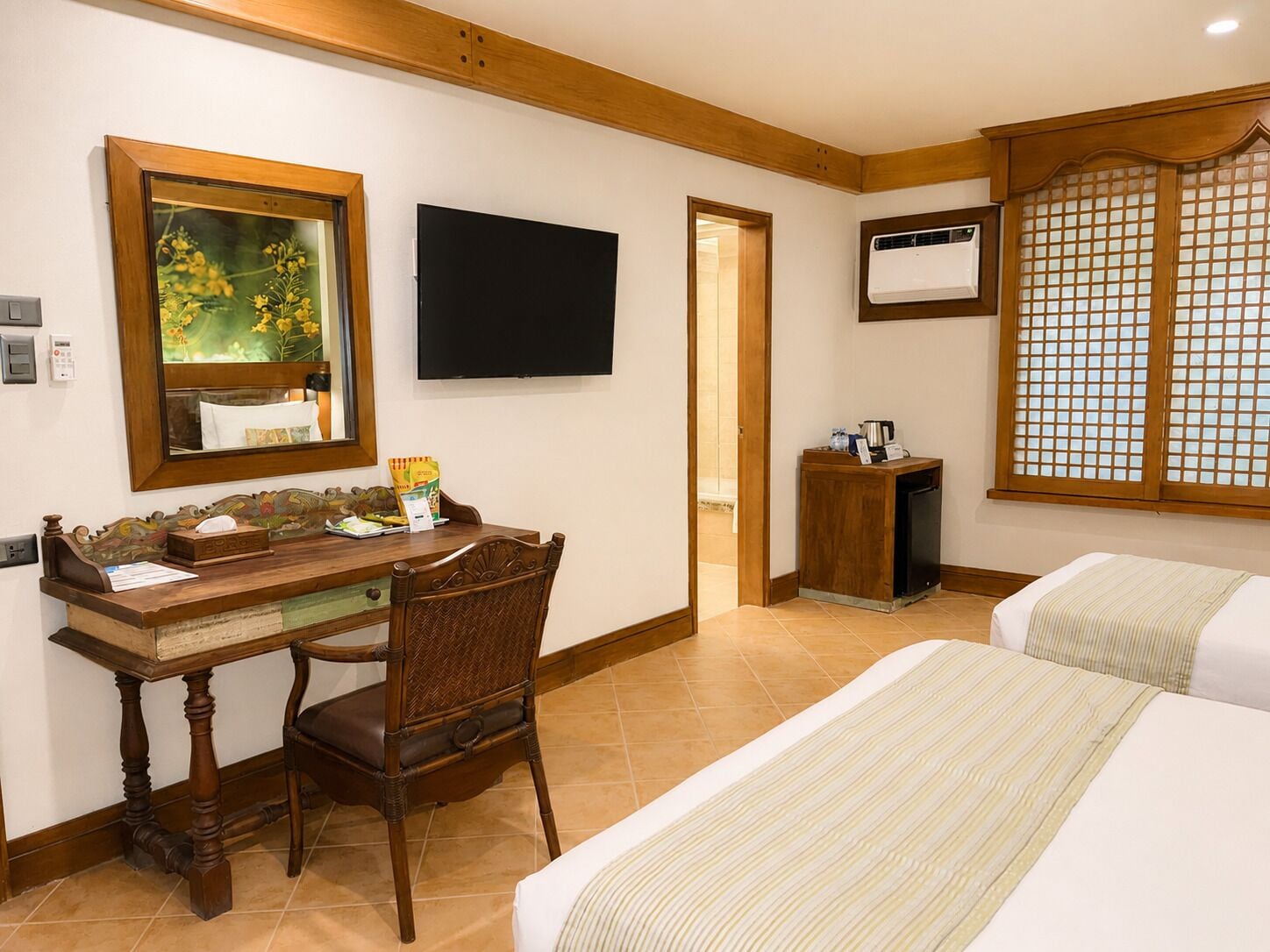 A hotel room with a wooden desk, a chair, a bed, a television, an air conditioning unit, and a window with a wooden frame.