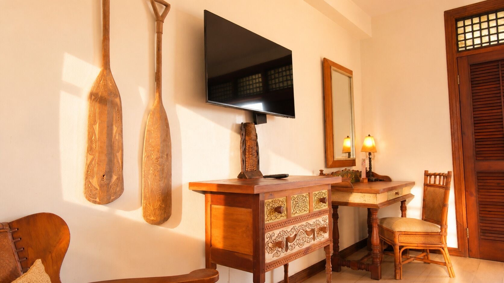 A room with a flat-screen TV mounted on the wall, a wooden cabinet with intricate designs, a chair, and a wooden door with a window.